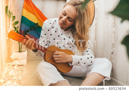 Young millennial hippie woman sitting on balcony play guitar. Music lesson and singer sitting. LGBTQ rainbow flag on background. Online music tuition. Playing instrument and enjoying songs and leisure 80523634