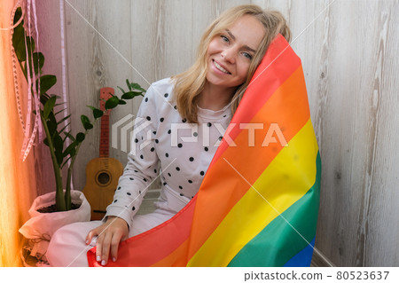 Young blonde millennial hippie woman with rainbow LGBTQ flag at home balcony. Peace and freedom the symbol of LGBT, spending and sharing loving time. Support LGBTQ community. 80523637