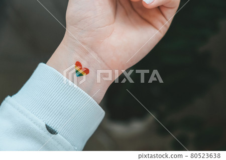 Closeup of young caucasian millennial hippie woman with a rainbow flag in heart shape painted in wrist. Rainbow color tattoo, symbol of LGBTQ pride month. Equal rights Closeup of young caucasian millennial hippie woman with a rainbow flag in heart shape painted in wrist. Rainbow color tattoo, symbol of LGBTQ pride month. Equal rights 80523638