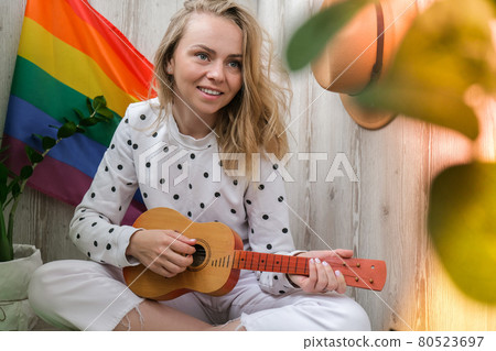 Young millennial hippie woman sitting on balcony play guitar. Music lesson and singer sitting. LGBTQ rainbow flag on background. Online music tuition. Playing instrument and enjoying songs and leisure 80523697