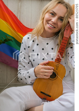 Young millennial hippie woman sitting on balcony play guitar. Music lesson and singer sitting. LGBTQ rainbow flag on background. Online music tuition. Playing instrument and enjoying songs and leisure 80523705