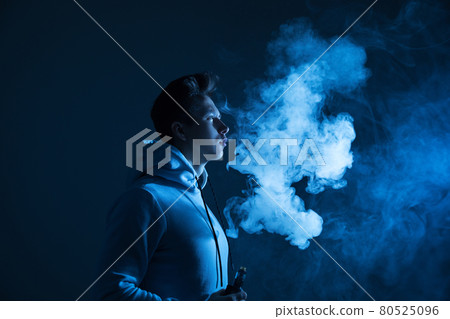 A young guy smokes a vape in the studio in neon light. A young guy smokes a vape in the studio in neon light. 80525096