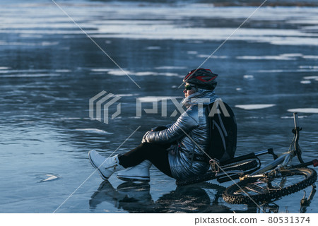 Woman sitting near her bike on ice. The girl cyclist stopped to rest. She sits on the wheel and enjoys the beautiful view of the sunset. Woman sitting near her bike on ice. The girl cyclist stopped to rest. She sits on the wheel and enjoys the beautiful view of the sunset. 80531374
