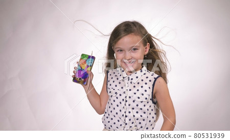 Child with headphones dancing at studio background. Girl listens to music on smartphone. Beautiful cute happy little girl listens to music. Child with headphones dancing at studio background. Girl listens to music on smartphone. Beautiful cute happy little girl listens to music. 80531939
