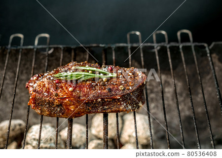 Hot spicy steak grilling on a summer barbecue over the hot coals garnished decorated with a branch of rosemary 80536408