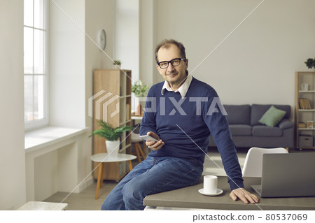Mature businessman with phone sitting on table with laptop in home office Mature businessman with phone sitting on table with laptop in home office 80537069