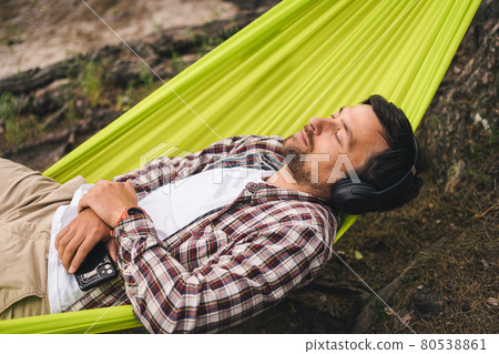 Man on bicycle trip at camping by lake is relaxing in green hammock while listening to music. Active recreation theme in nature. Hipster cyclist with headphones having fun in hammock by river Man on bicycle trip at camping by lake is relaxing in green hammock while listening to music. Active recreation theme in nature. Hipster cyclist with headphones having fun in hammock by river 80538861
