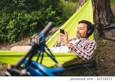 Man travels on bicycle, relaxing in green hammock, surfing Internet on smartphone, listening music on headphones in forest near lake. Cyclist in hammock at campsite by river. Male on bike in hammock 80538872