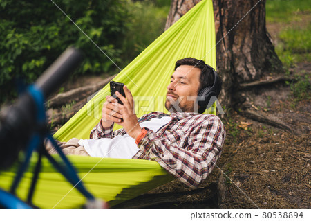 Outdoors and adventure concept. Bike trip to forest. Cyclist is resting in green hammock between trees in nature by lake while listening to music on headphones and surfing Internet on smartphone 80538894