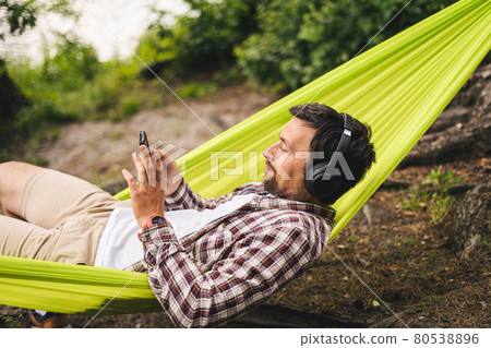 Young caucasian man resting in hammock, listening to music on headphones and using smartphone afterwards on bicycle in forest near the lake. Audio healing. Idyllic place. Travel, camping in nature 80538896