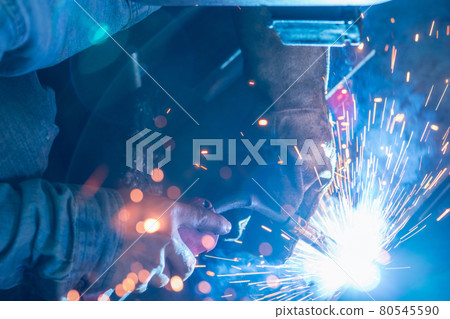 Welder welding metal with argon arc welding machine and has welding sparks. A man wears welding mask and protective gloves. Safety in industrial workplace. Welder working with safety. Steel industry. 80545590