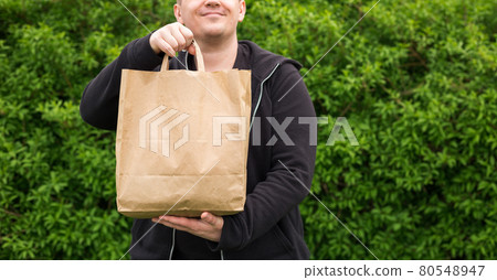Close up of man hand with eco paper bag for takeaway food on nature green background. Delivery in any weather around the clock to the client. Close up of man hand with eco paper bag for takeaway food on nature green background. Delivery in any weather around the clock to the client. 80548947