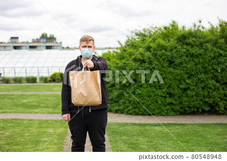 Close up of man hand in mask holding paper bag for takeaway food on nature green background. Delivery in any weather around the clock to the client. 80548948