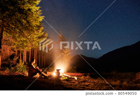 Female traveler sitting near campfire under night starry sky. Female traveler sitting near campfire under night starry sky. 80549050