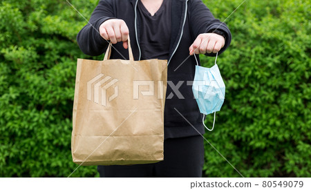 Close up of man hand with mask and paper bag for takeaway food on nature green background. Delivery in any weather around the clock to the client. Close up of man hand with mask and paper bag for takeaway food on nature green background. Delivery in any weather around the clock to the client. 80549079