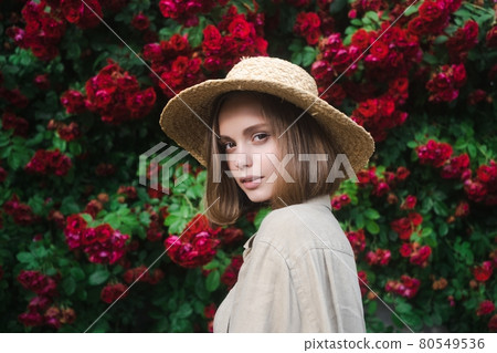 Elegant beautiful blonde young woman in a straw hat on the background of a bush of blooming roses. 80549536