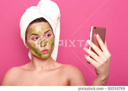 Emotional adorable cosmetologist making selfie for social networking sites, holding smartphone in her hand, head wrapped in towel, posing half naked, having facial green mask applied on skin. Emotional adorable cosmetologist making selfie for social networking sites, holding smartphone in her hand, head wrapped in towel, posing half naked, having facial green mask applied on skin. 80550769