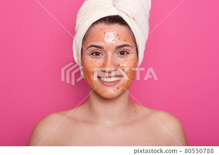 Horizontalshot of beautiful girl wrapped hair with white towel, skin covered by mask. Model with mask on face, posing with bare shoulders, standig indoor against pink studio wall. Beauty concept. Horizontalshot of beautiful girl wrapped hair with white towel, skin covered by mask. Model with mask on face, posing with bare shoulders, standig indoor against pink studio wall. Beauty concept. 80550788