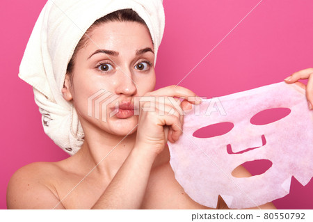 Portrait of half naked cute young woman with white towel on her head, looking directly at camera, showing face cosmetic mask after applying it, face revitalizing procedure, looking surprised. 80550792