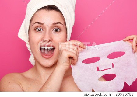 Headshot of amazed beautiful young woman holds cosmetic mask, feels refreshed and energized, has wrapped towel on head, looks with amazement directly at camera, isolated over pink background Headshot of amazed beautiful young woman holds cosmetic mask, feels refreshed and energized, has wrapped towel on head, looks with amazement directly at camera, isolated over pink background 80550795