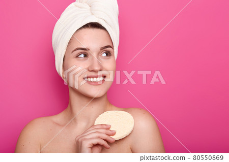 Positive optimistic young woman wipes body with bath sponge, has toothy smile, shows white teeth, looks away, shows bare shoulders, has white towel wrapped on head, isolated over pink background Positive optimistic young woman wipes body with bath sponge, has toothy smile, shows white teeth, looks away, shows bare shoulders, has white towel wrapped on head, isolated over pink background 80550869