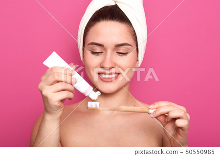 Indoor shot of beautiful attentive lady squeezing toothpaste from tube on her wooden toothbrush, doing hygiene procedures, smiling sincerely, standing naked, covering her hair with white towel. Indoor shot of beautiful attentive lady squeezing toothpaste from tube on her wooden toothbrush, doing hygiene procedures, smiling sincerely, standing naked, covering her hair with white towel. 80550985