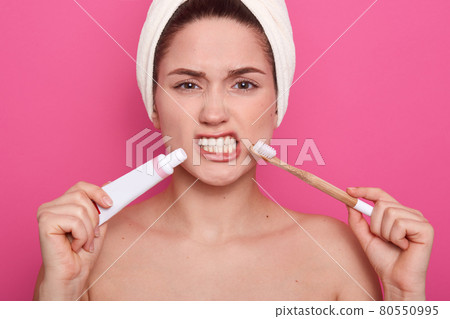 Image of attractive woman with dark hair, holding toothbrushe and tooth paste in hands, has angry facial expression, female looking at camera, posing with bere shoulders and white towel on head. 80550995