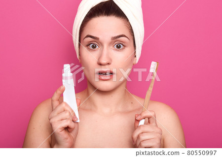 Woman brushing her teeth with shocked facial expression, girl with toothbrush isolated on pink studio background, lady with white towel on head, holds toothpaste and toothbrush in hands. Oral hygiene. Woman brushing her teeth with shocked facial expression, girl with toothbrush isolated on pink studio background, lady with white towel on head, holds toothpaste and toothbrush in hands. Oral hygiene. 80550997