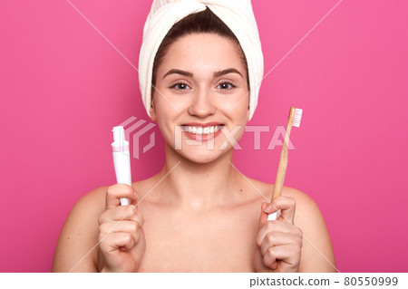Good looking charming lady smiling sincerely, holding tube of whitening toothpaste and wooden toothbrush in both hands, looking directly in camera, being in high spirits. Dental care concept. Good looking charming lady smiling sincerely, holding tube of whitening toothpaste and wooden toothbrush in both hands, looking directly in camera, being in high spirits. Dental care concept. 80550999