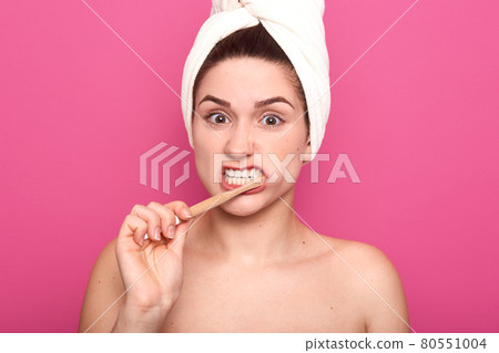 Indoor studio shot of busy unsatisfied young female brushing her teeth with efforts, having unpleasant facial expression, covering her hair with white towel, being naked. Dental care concept. Indoor studio shot of busy unsatisfied young female brushing her teeth with efforts, having unpleasant facial expression, covering her hair with white towel, being naked. Dental care concept. 80551004