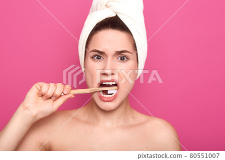 Indoor studio shot of displeased energetic young woman looking directly at camera, brushing teeth intesively with toothbrush, being naked with towel on head, standing isolated over pink background. Indoor studio shot of displeased energetic young woman looking directly at camera, brushing teeth intesively with toothbrush, being naked with towel on head, standing isolated over pink background. 80551007