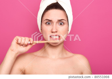 Closeup portrait of emotional unsatisfied woman with white towel on her head, brushing her teeth with professional equipment, having unpleasant facial expression, opening her eyes widely with terror. Closeup portrait of emotional unsatisfied woman with white towel on her head, brushing her teeth with professional equipment, having unpleasant facial expression, opening her eyes widely with terror. 80551008