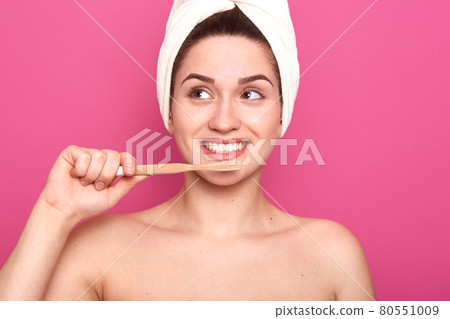 Young cheerful woman with perfect white smile looking aside, holding wooden toothbrush in front of face, being naked, having smooth well kept skin, posing isolated over pink background in studio. Young cheerful woman with perfect white smile looking aside, holding wooden toothbrush in front of face, being naked, having smooth well kept skin, posing isolated over pink background in studio. 80551009