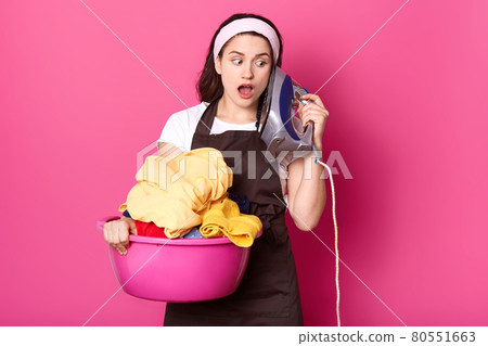 Close op portrait of cute good looking young woman with surprised facial expression, holds pink basin full of clean clothes and keeps iron like phone, imagine conversation, has fun while ironig. 80551663
