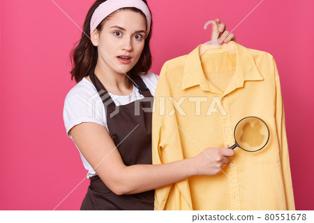 Young woman with dirty clothes isolated over pink background, being shocked, holding magnifier in front of spot, looks at camera, needs to wash yellow shirt. 80551678