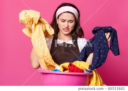 Disappointed emotional young lady wearing white headband and t shirt, brown apron, having unpleasant facial expression, sorting out dirty clothes, looks helpless. People and house chores concept. 80551681
