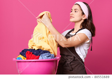 Profile of delighted attractive brunette holding light shirt after washing, being satisfied with result, smiling sincerely, posing isolated over pink background in studio. Housewife lifestyle concept. Profile of delighted attractive brunette holding light shirt after washing, being satisfied with result, smiling sincerely, posing isolated over pink background in studio. Housewife lifestyle concept. 80551689