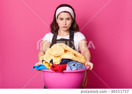 Dark haired woman fastidiously looks at dirty linen in basin. Young housewife does laundry. Dirty cream colerd towel. Girl with white hair band and brown apron helps about house. Housekeeping concept. Dark haired woman fastidiously looks at dirty linen in basin. Young housewife does laundry. Dirty cream colerd towel. Girl with white hair band and brown apron helps about house. Housekeeping concept. 80551737