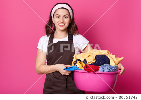 Young smiling beautiful female walking with pink basin full of dirty items of clothes, holding it with both hands, looks positive. Busy attractive housewife stands isolated over pink background. 80551742