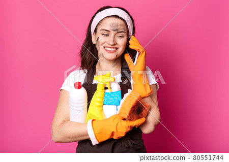 Positive Caucasian woman speaking on mobile phone while clening her house, holding sponge and detergents in hands, dressed in casual clothes, wears rubber gloves and apron, isolated over pink wall. Positive Caucasian woman speaking on mobile phone while clening her house, holding sponge and detergents in hands, dressed in casual clothes, wears rubber gloves and apron, isolated over pink wall. 80551744