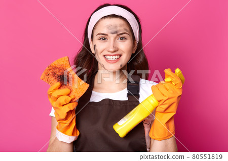 Enthusiastic smiling housewife holds dirty sponge and cleaning detergent, satisfied with result of cleaning, being dirty. Charismatic brunette female wears brown apron, t shirt and headband. 80551819