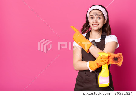 Enthusiastic cheerful woman standing isolated over pink background in studio, holding detergent and dirty washcloth in one hand, showing direction with forefinger, looking directly at camera. Enthusiastic cheerful woman standing isolated over pink background in studio, holding detergent and dirty washcloth in one hand, showing direction with forefinger, looking directly at camera. 80551824
