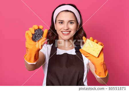Indoor shot of charming young Caucasian housewife wearing headband, yellow rubber gloves and brown apron, holding sponges in hands, having posintive expression, looking smiling directly at camera. Indoor shot of charming young Caucasian housewife wearing headband, yellow rubber gloves and brown apron, holding sponges in hands, having posintive expression, looking smiling directly at camera. 80551871