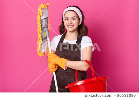 Smiling female wears brown apron and yellow rubber gloves, holds mop and red bucket. Young woman cleans house. Beautiful girl does housework. Attractive housewife works at home. Household cocept. Smiling female wears brown apron and yellow rubber gloves, holds mop and red bucket. Young woman cleans house. Beautiful girl does housework. Attractive housewife works at home. Household cocept. 80551880