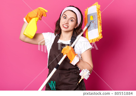 Close up of young fun housewife in orange gloves, brown apron, white t shirt, hair band. Housekeeper woman shoots from spray bottle with cleaner liquid, sick and tired of doing chores. Hygiene concept 80551884