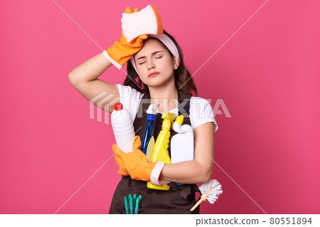 Indoor shot of young tired housewife wearing brown apron, white casual t shirt, haedband, posing with cleaning deretgents isolated over pink background, maid touching her forehead, holds sponge. Indoor shot of young tired housewife wearing brown apron, white casual t shirt, haedband, posing with cleaning deretgents isolated over pink background, maid touching her forehead, holds sponge. 80551894
