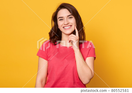 Cheerful young Caucasian woman with gentle charming smile, wearing red shirt, having pleasant appearance, keeps index finger on cheek, waits kiiss, smiles happily, poses against yellow background. Cheerful young Caucasian woman with gentle charming smile, wearing red shirt, having pleasant appearance, keeps index finger on cheek, waits kiiss, smiles happily, poses against yellow background. 80552581