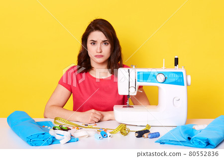 Close up portrait of sad seamstress working at her workshop, adorable dressmaker wearing red casual t shirt, surrounded different equipments, looks upset, has mistake, isolated over yellow background. Close up portrait of sad seamstress working at her workshop, adorable dressmaker wearing red casual t shirt, surrounded different equipments, looks upset, has mistake, isolated over yellow background. 80552836