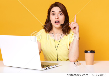Half lengh photo of office worker sitting with opened mouth and eyes, sitting with raised forefinger, isolated over yellow background in studio, having new idea, looks astonished, online manager. 80553038