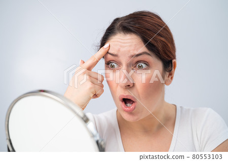 Caucasian woman looks at herself in the mirror and is upset because of facial wrinkles on her face. Caucasian woman looks at herself in the mirror and is upset because of facial wrinkles on her face. 80553103